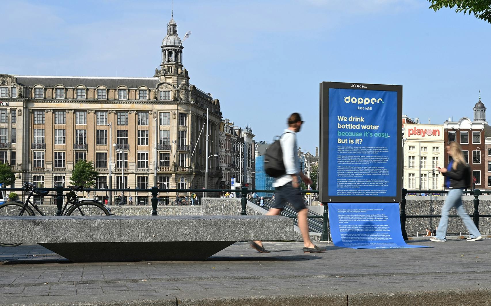 Two people walk past a big blue Dopper Abri campaign at Amsterdam Central